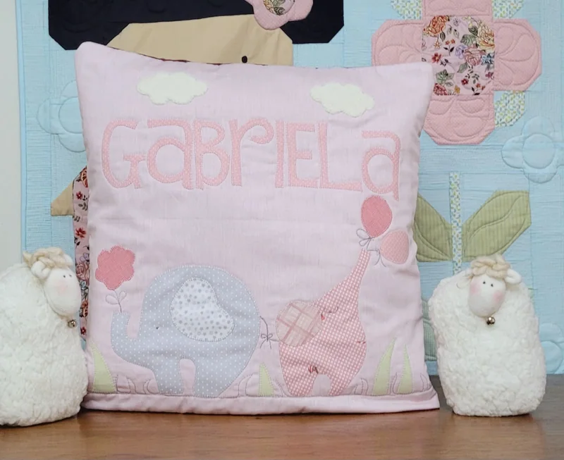 Almofada rosa com o nome Gabriela, elefante, girafa e balão aplicados, entre duas ovelhinhas de tecido.