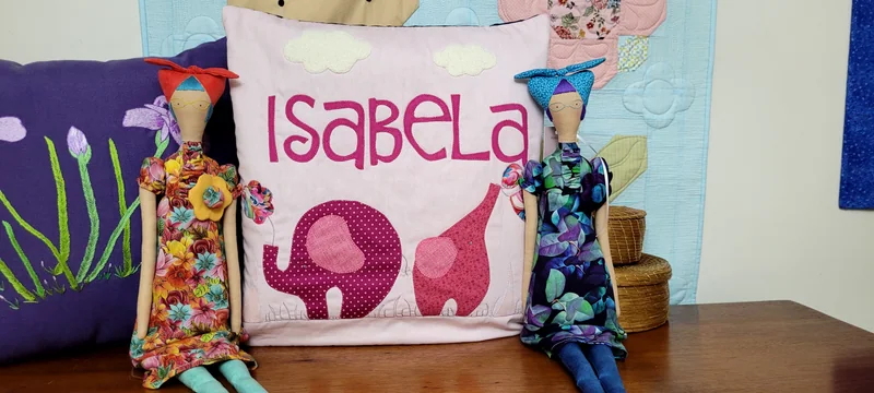 Almofada rosa com o nome Isabela, elefante e girafa aplicados, entre bonecas de vestidos estampados.