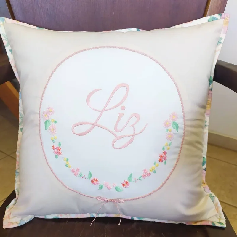 Almofada branca com o nome Liz bordado em rosa e um círculo de pequenas flores ao redor.