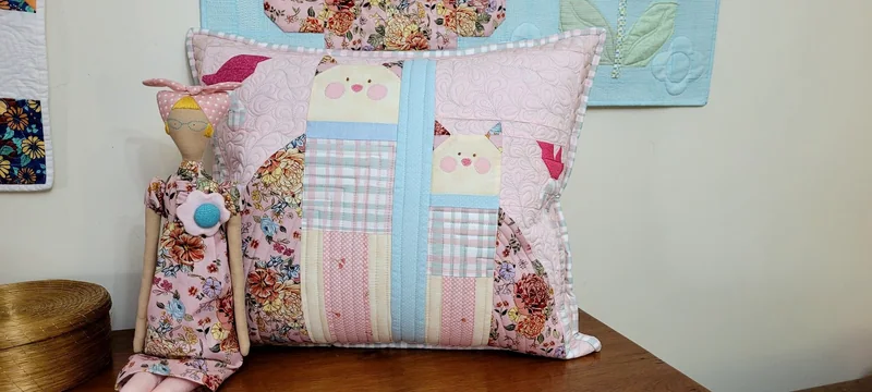 Almofada de retalhos rosa, azuis e florais, acompanhada de uma boneca de tecido.
