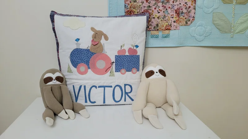 Almofada clara com o nome Victor e aplicações coloridas, entre bichinhos de tecido.
