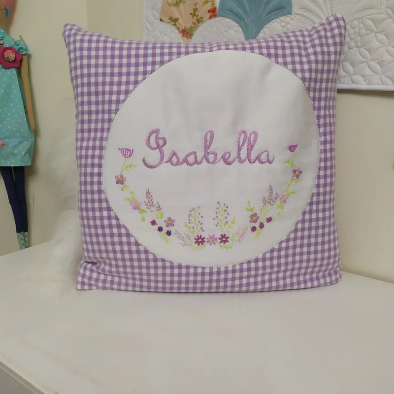 Almofada com o nome Isabella e flores bordadas, cercada de tecido xadrez lilás.