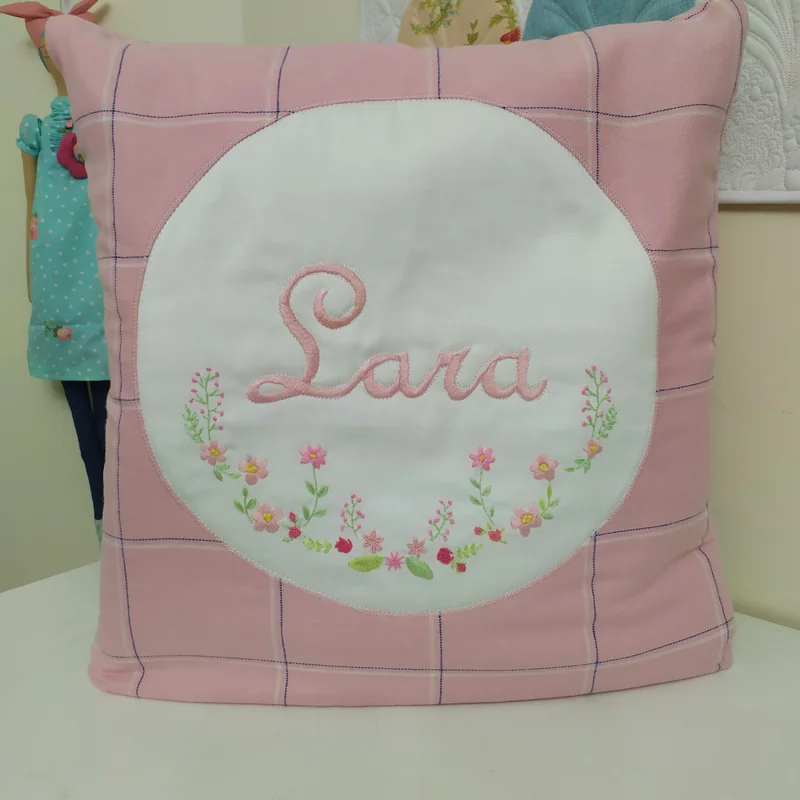 Almofada rosa com o nome Lara e pequenos ramos bordados sobre um círculo claro.