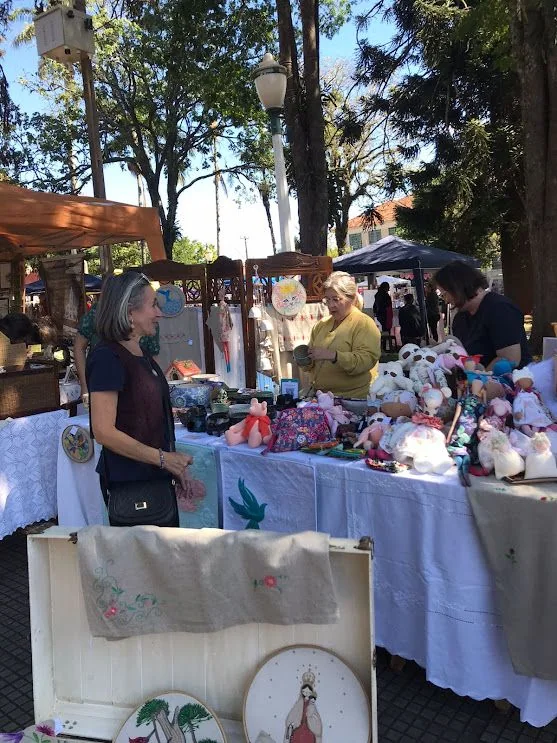 Pessoas ao redor de mesas com trabalhos artesanais em um bazar ao ar livre.