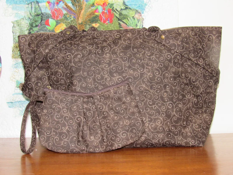Bolsa marrom com arabescos claros e alça comprida, ao lado de uma peça menor.