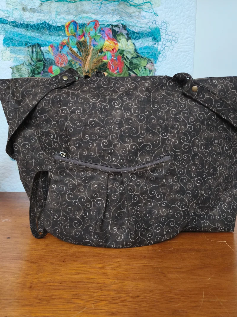 Bolsa escura de tecido com arabescos e alças compridas, sobre a mesa.
