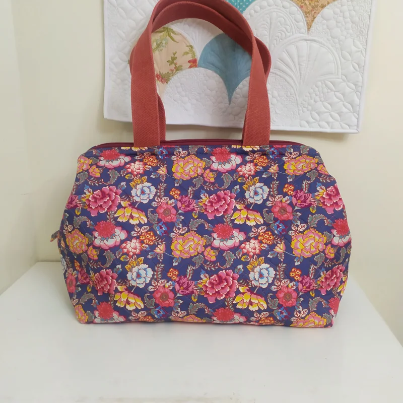 Bolsa de flores coloridas sobre fundo azul-escuro, com alças vermelhas, apoiada em uma cômoda branca.