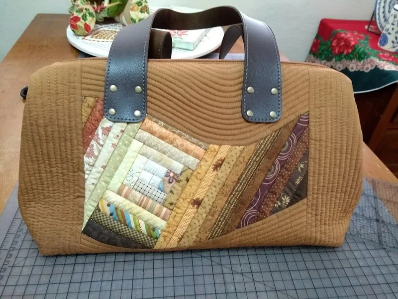 Bolsa ocre com painel geométrico de patchwork e alças escuras.