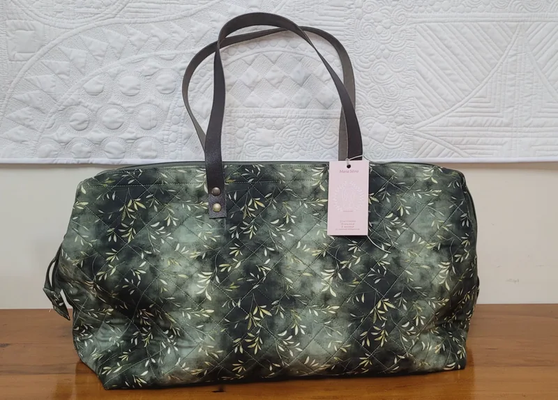 Bolsa de viagem verde-musgo com ramos claros, alças cinza e etiqueta do atelier.