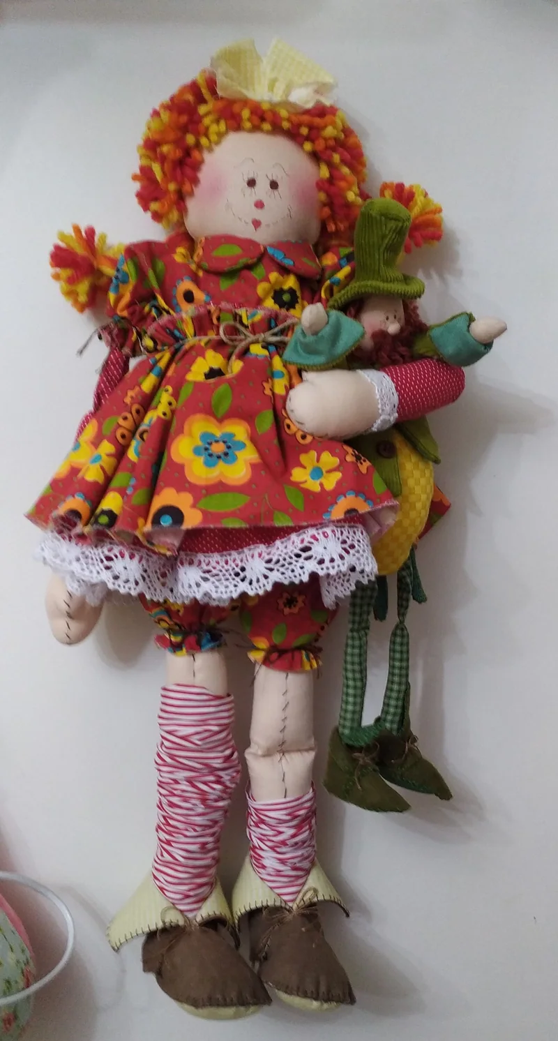 Boneca pendurada na parede, com tranças ruivas, vestido vermelho de flores, meias em rosa e branco e um pequeno pássaro verde na mão.