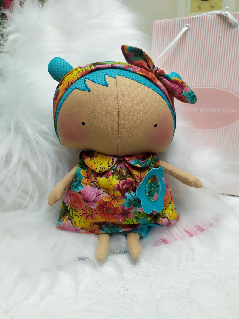 Boneca sentada sobre tecido felpudo, com cabelo azul, laço florido na cabeça e vestido de flores em rosa, amarelo e azul.