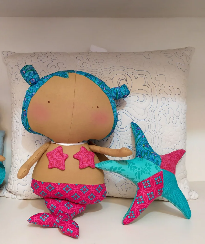 Boneca sereia de cabelo azul e cauda estampada em rosa e azul, encostada em uma almofada clara, ao lado de uma estrela-do-mar azul.