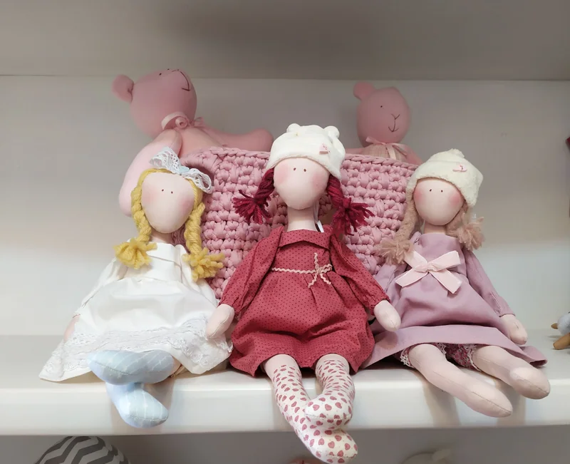Três bonecas sentadas, com toucas claras e vestidos branco, vinho e rosa.