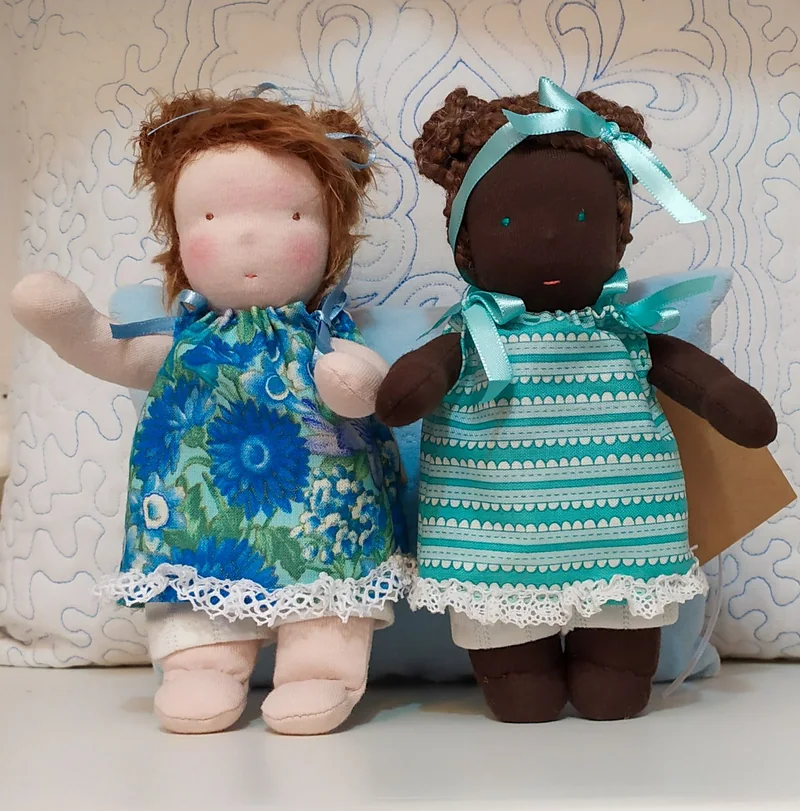 Duas bonecas, uma ruiva de vestido floral azul e outra de pele marrom com vestido listrado em verde-água, ambas com laços azuis.