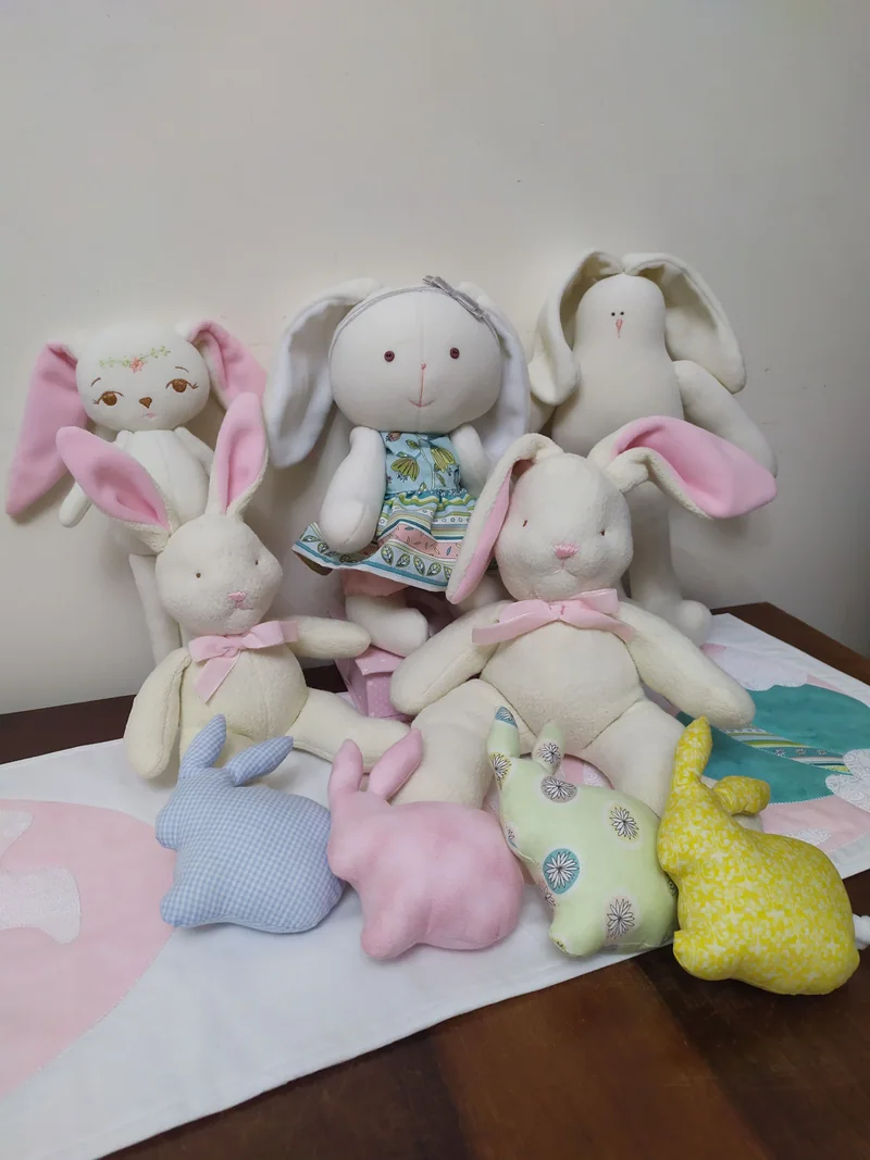 Coelhos claros de orelhas rosa e pequenos coelhinhos em lilás, azul, rosa, verde e amarelo, sobre uma manta com balão aplicado.