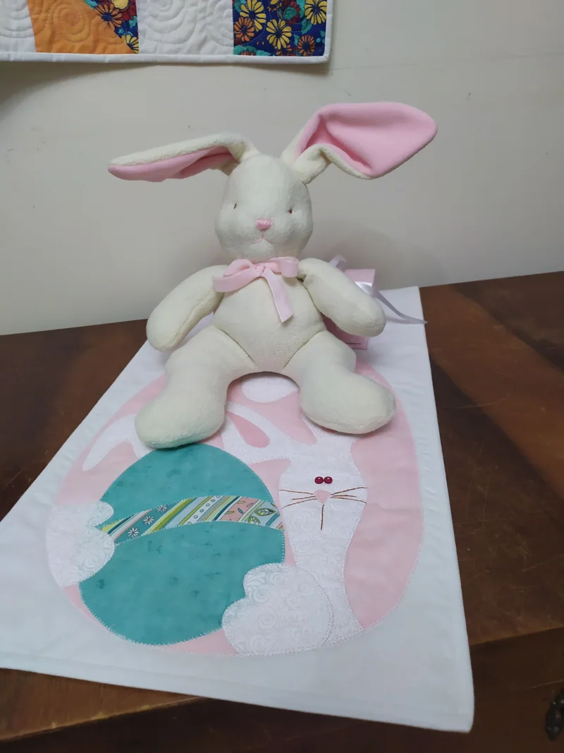 Coelho de tecido cru com orelhas rosa deitado sobre a manta clara, que traz um balão verde e rosa aplicado.