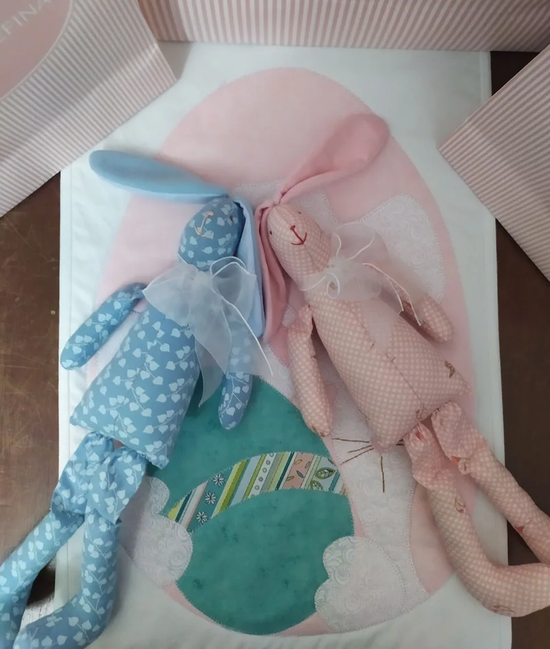 Dois coelhos compridos, um azul de bolinhas e outro rosa, deitados sobre a manta e junto das embalagens do atelier.