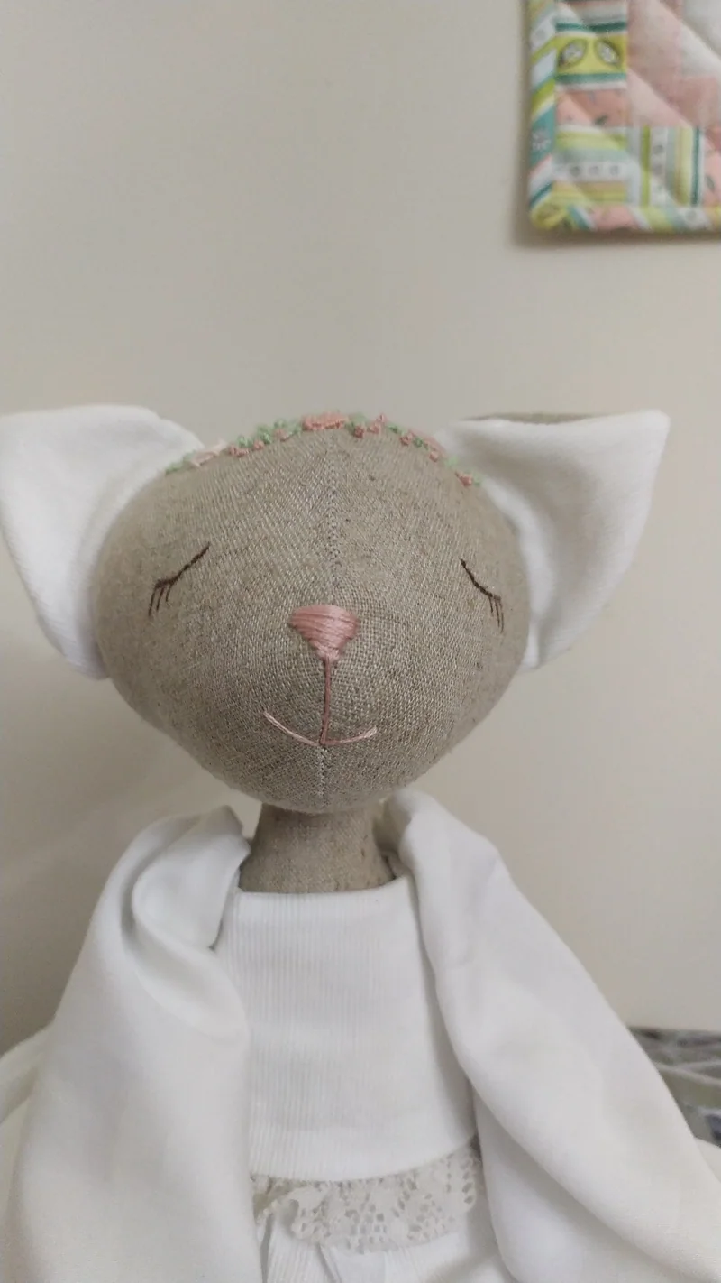 Gatinha de tecido em tom cru, com roupa branca e olhos fechados, diante de uma parede clara.