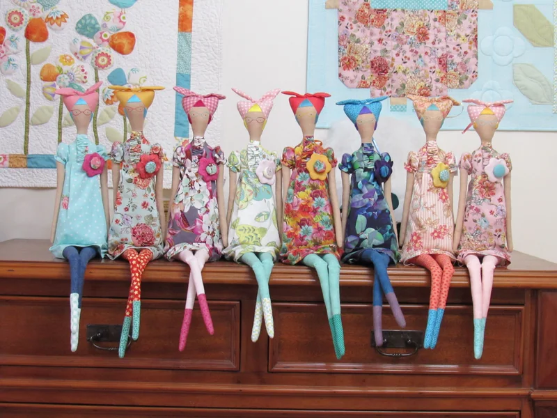 Sete gatos de tecido sentados lado a lado, com vestidos de diferentes estampas.