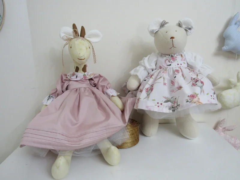 Girafa de vestido rosa e ursinha de vestido floral sentadas lado a lado.