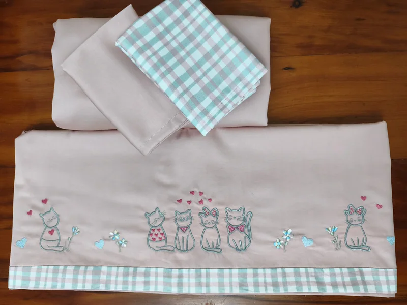 Lençol rosa-claro com uma fileira de gatinhos bordados e barra xadrez em verde e branco, com peças dobradas ao lado.