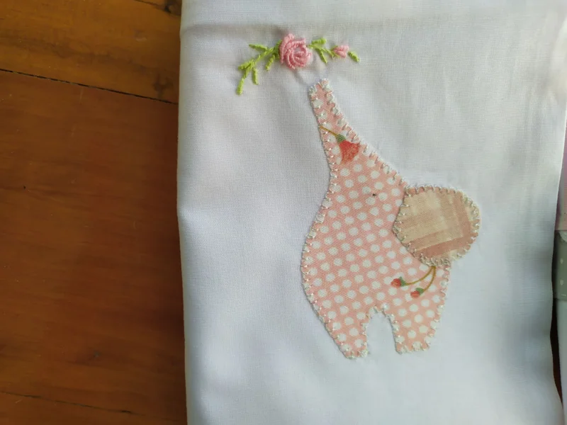 Detalhe de um lençol branco com girafa aplicada em xadrez rosa e flores bordadas na cabeça.