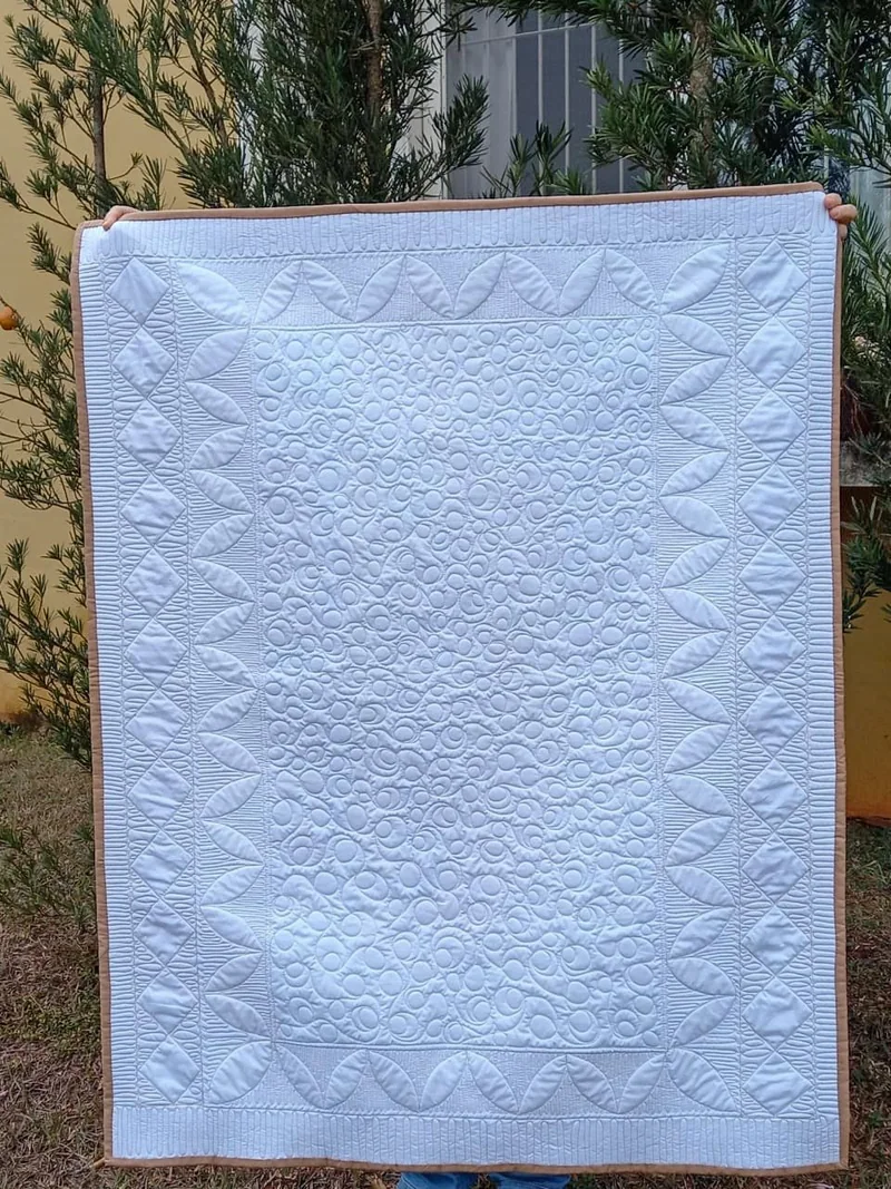 Manta branca fotografada ao ar livre, com o desenho do quilting visível nas bordas e no centro.