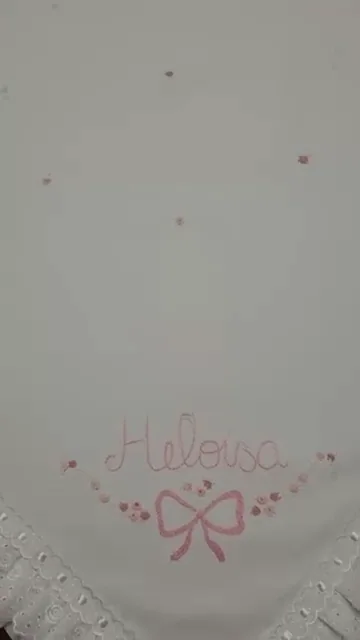 Detalhe do nome Heloísa e de um laço rosa bordados em uma manta clara.