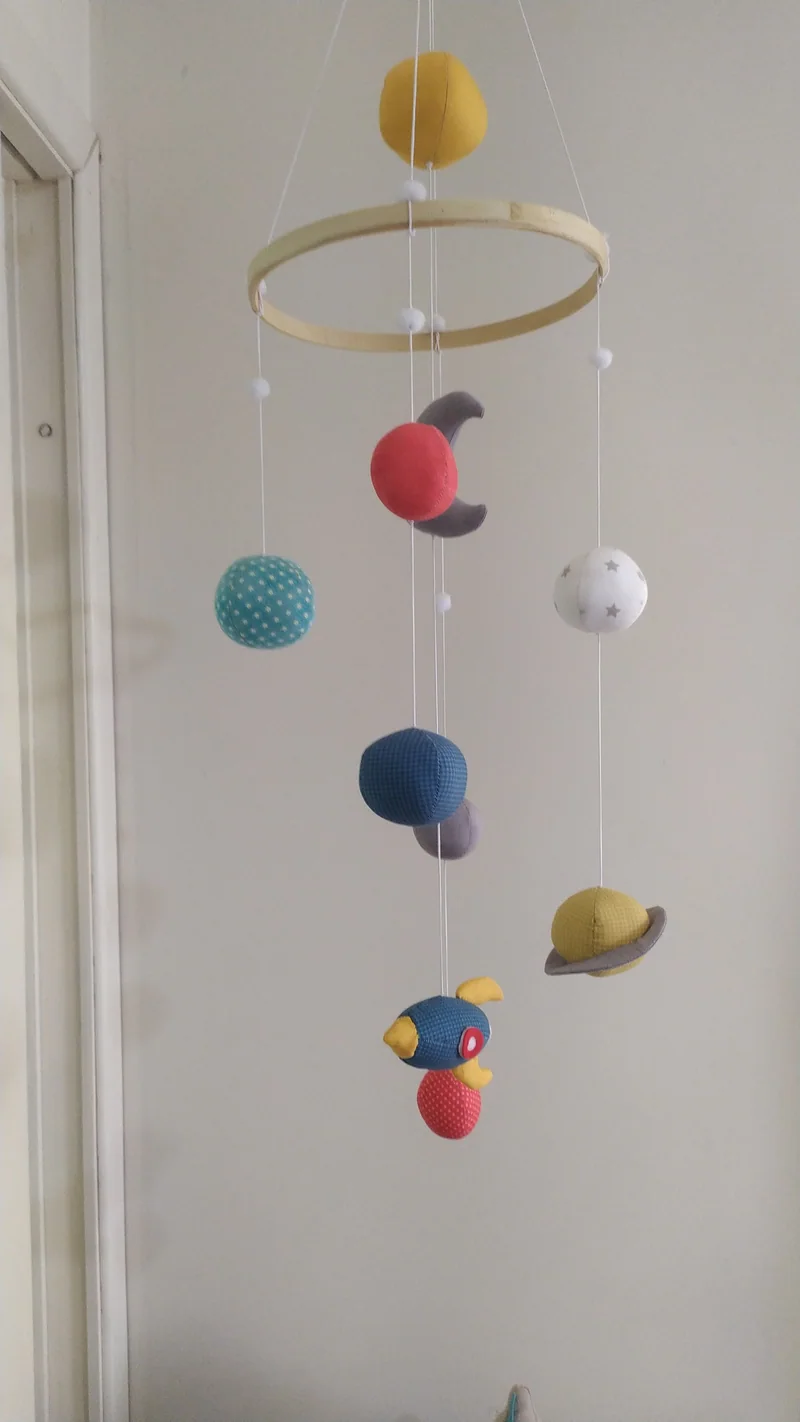 Esferas de tecido coloridas e pequenos pássaros pendurados em aro de madeira, diante de uma parede clara.