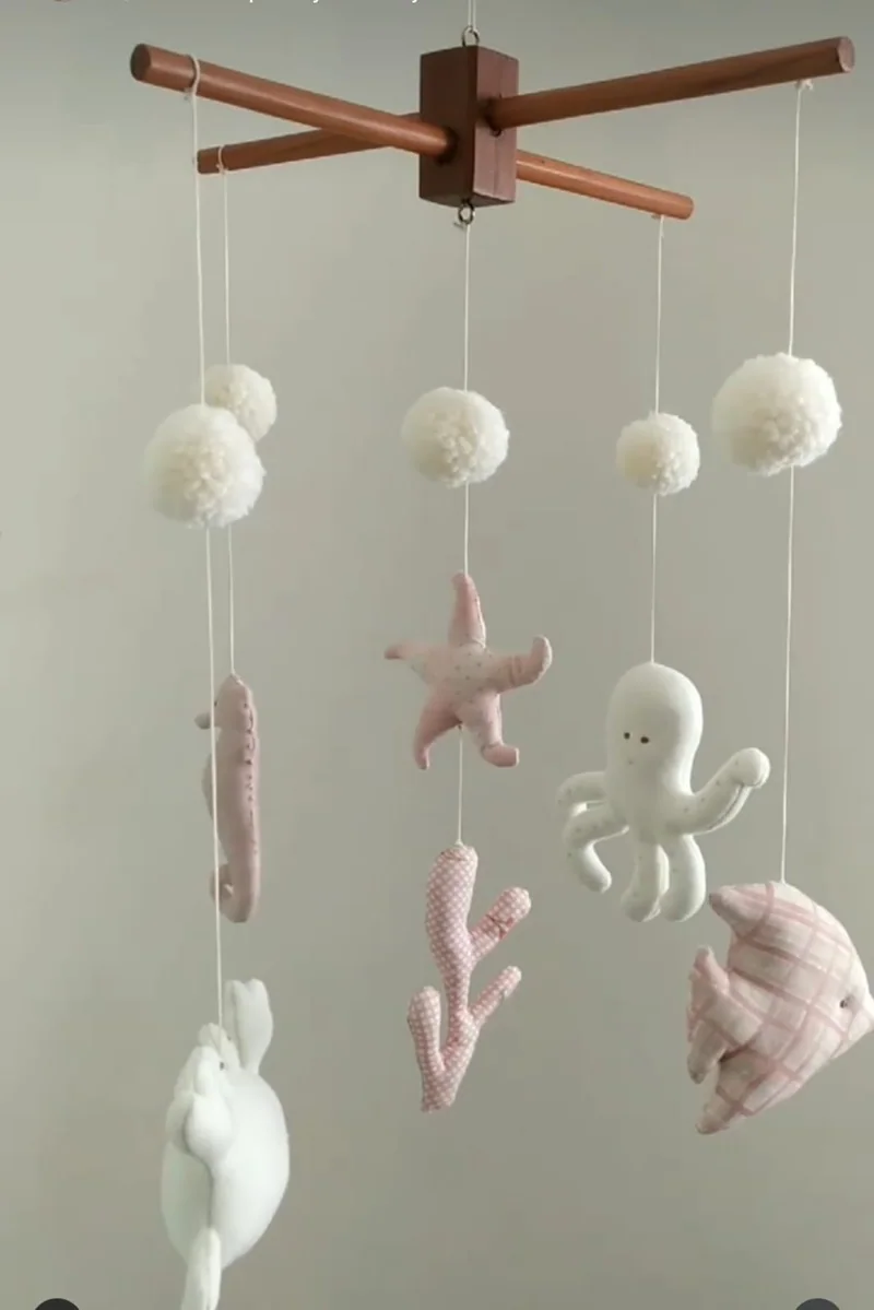 Suporte de madeira em cruz com pompons brancos e bichos do mar em rosa e branco pendurados abaixo.