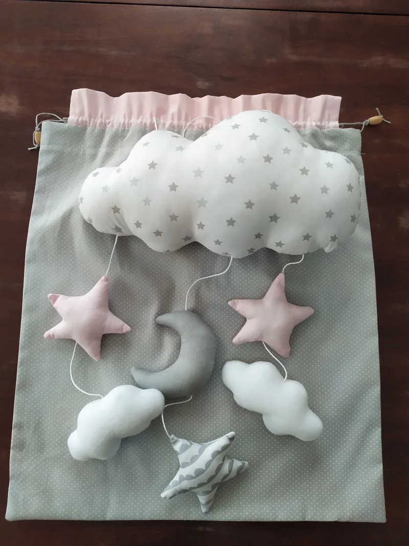 Nuvem cinza de estrelinhas, lua, estrelas rosa e nuvens brancas dispostas sobre um saco de tecido cinza com acabamento rosa.