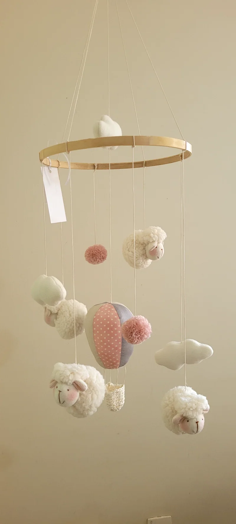 Ovelhinhas brancas, balões em rosa e coral, nuvens e pompons pendurados em aro de madeira, com a etiqueta do atelier.