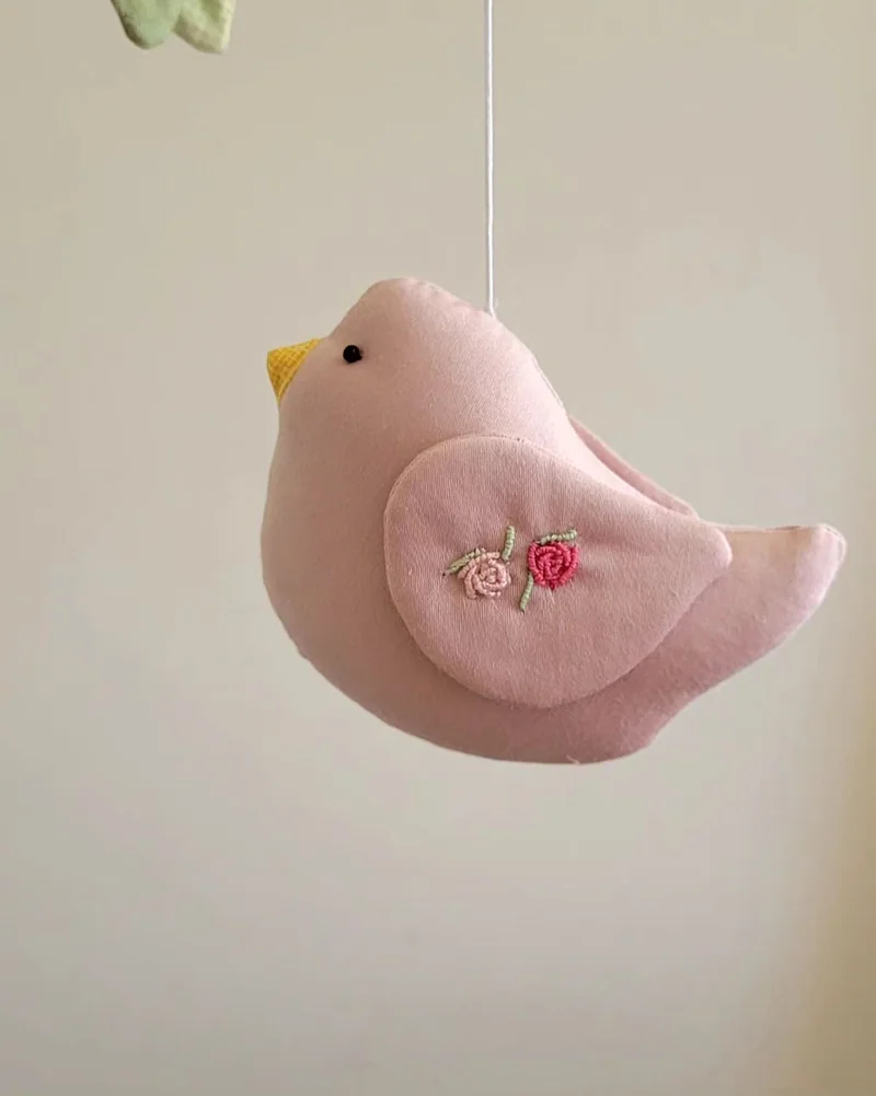 Detalhe de um pássaro rosa com uma flor bordada na asa.