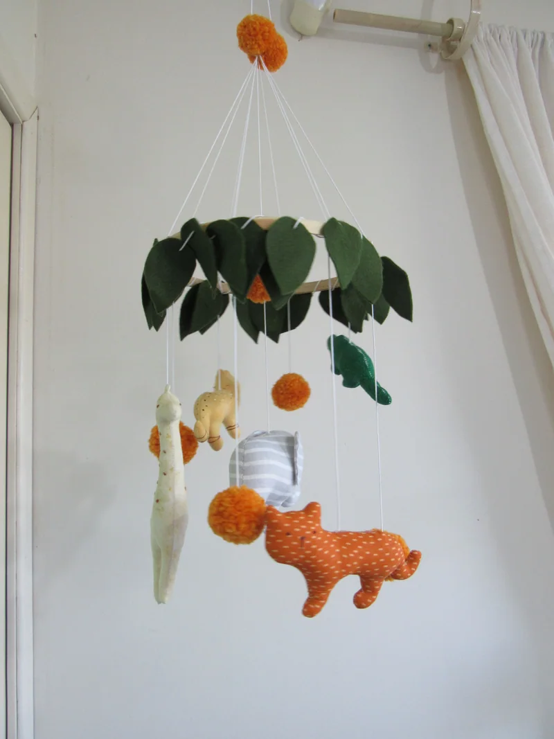 Móbile de safári com animais de tecido, folhas verdes e esferas laranja.