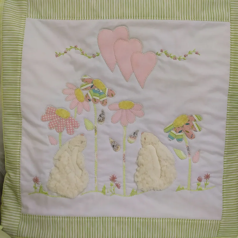 Painel com dois coelhos brancos aplicados entre flores rosa, corações e pássaros bordados, com borda listrada verde.