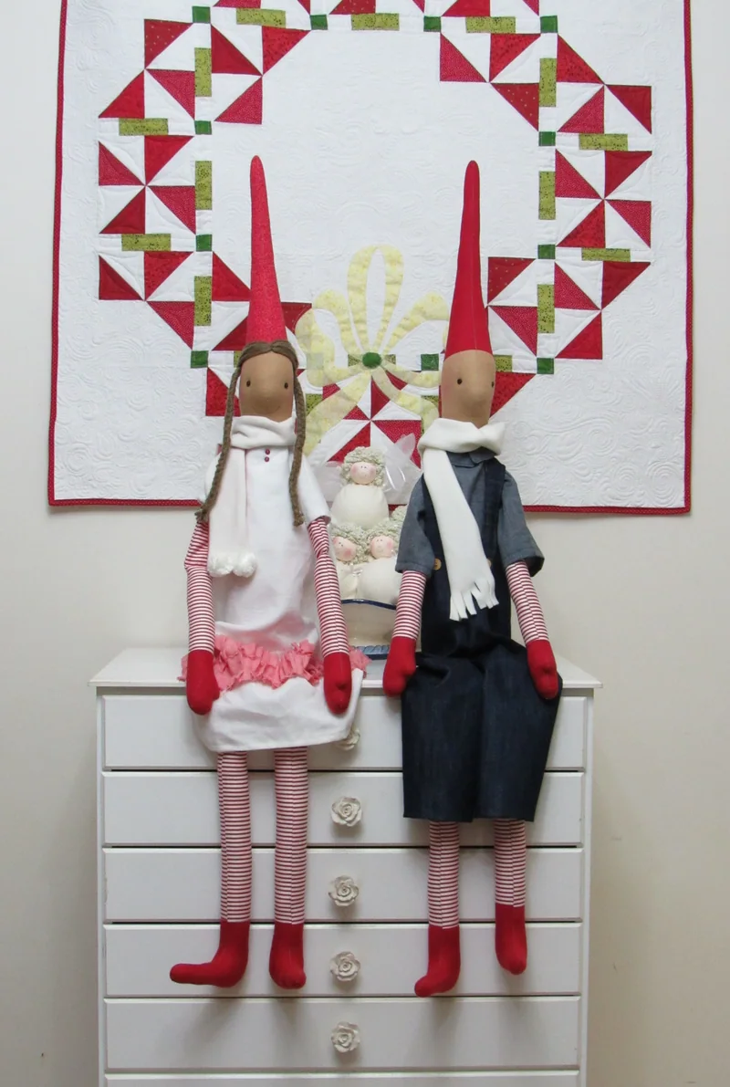 Bonecos de Natal de gorros compridos sentados diante de um painel de patchwork.