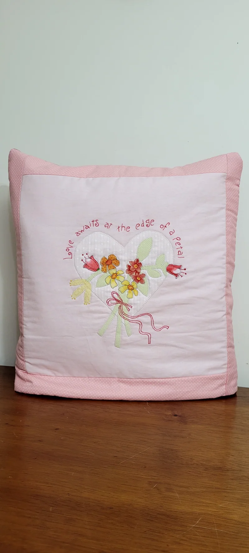 Almofada rosa com flores bordadas ao centro e bordas em rosa mais forte.