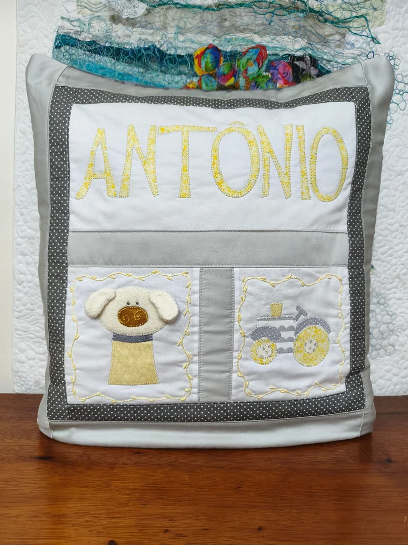 Almofada clara com o nome Antonio em amarelo, cachorro e trator aplicados, com molduras cinza.