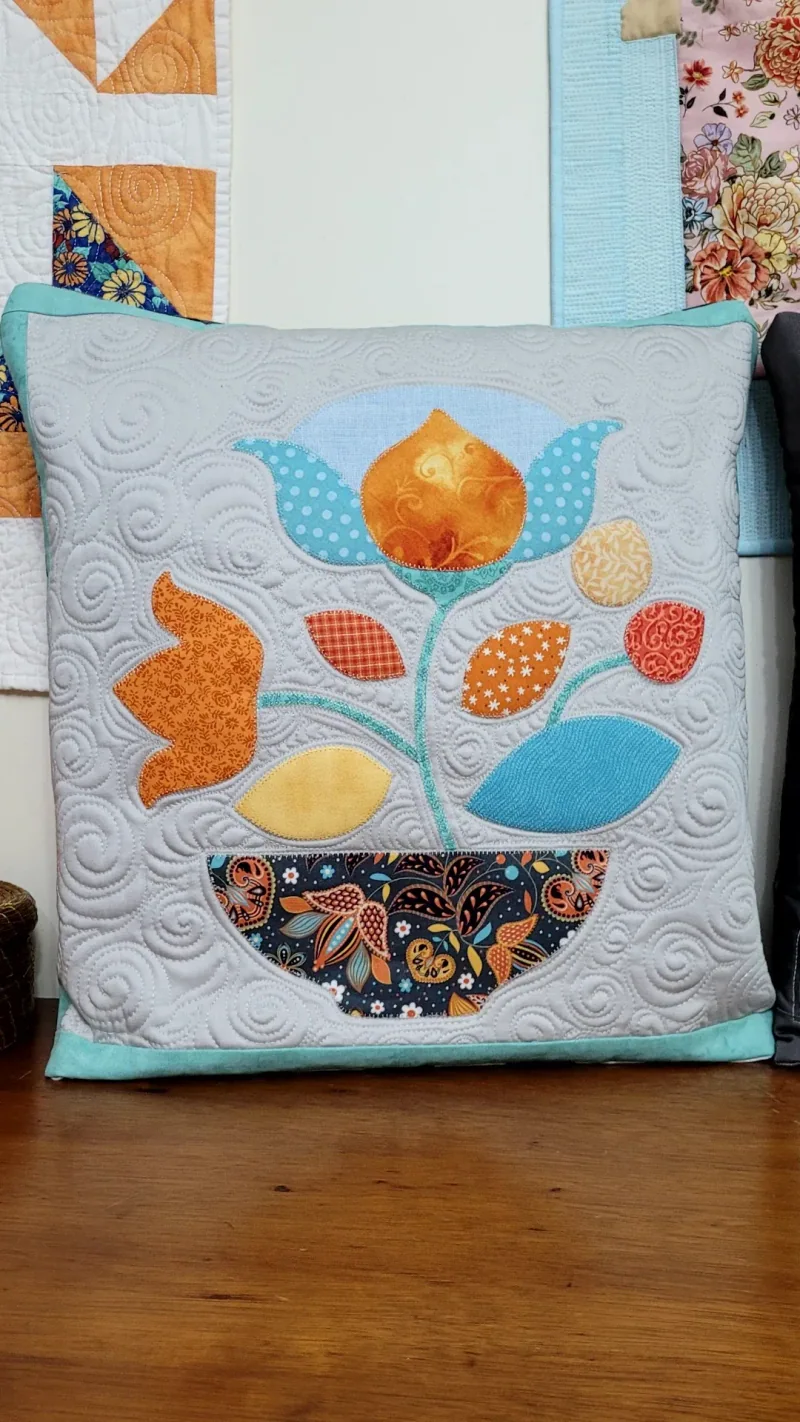 Almofada com flor laranja e azul aplicada, quilting ao redor e borda azul-clara.
