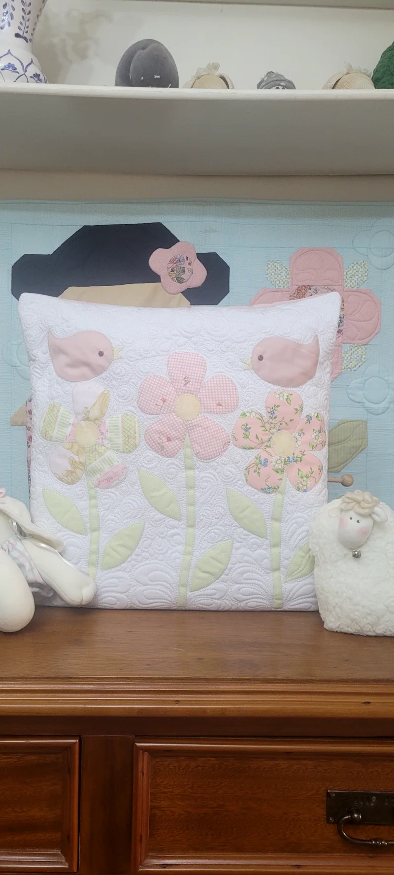 Almofada branca com flores em rosa e verde aplicadas e quilting, sobre a cômoda.