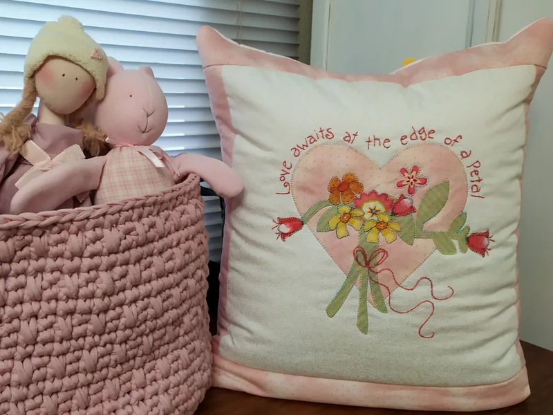 Almofada com coração de flores bordado, frase ao redor e bordas rosa, ao lado de uma cesta de bonecas.