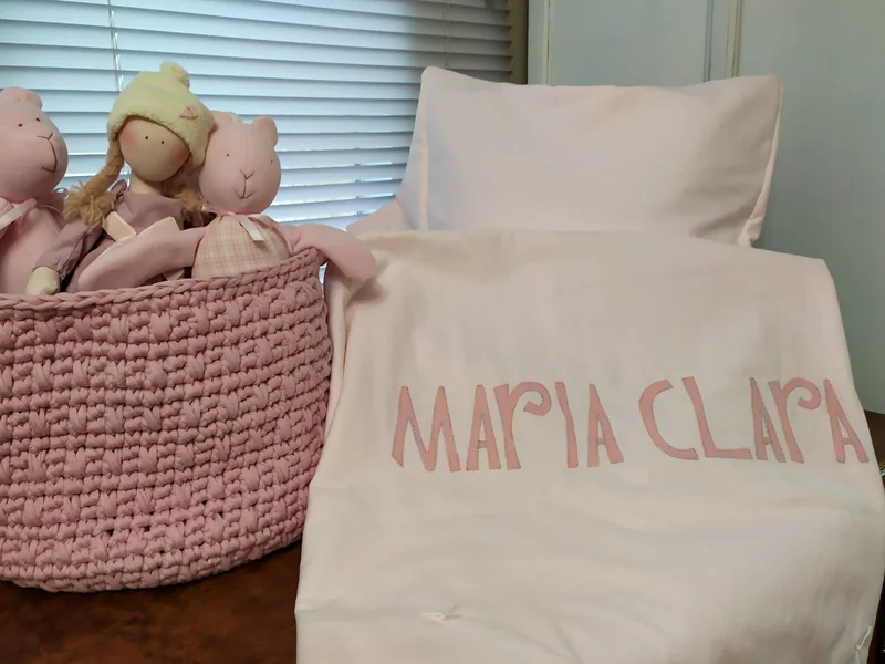 Manta rosa-clara com o nome Maria Clara bordado, ao lado da cesta de bonecas.