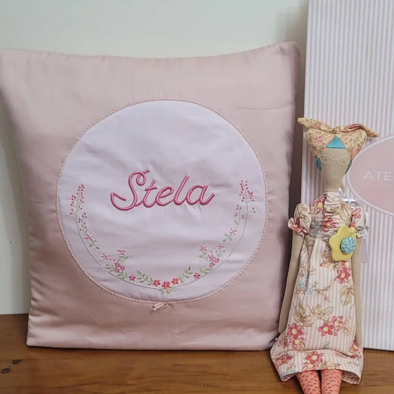 Quillow rosa com o nome Stela e flores bordadas, acompanhado de uma boneca.