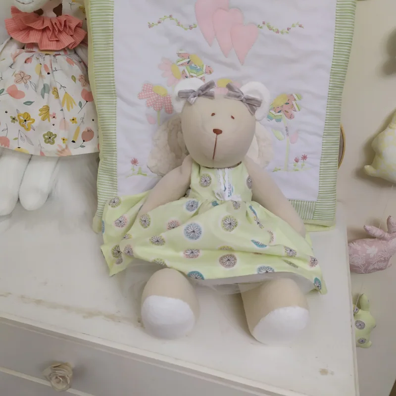 Ursa sentada em uma cômoda branca, com vestido amarelo florido, e um painel bordado com balão apoiado atrás.