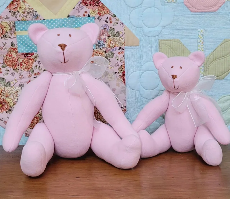 Duas ursinhas cor-de-rosa, em tamanhos diferentes, diante de uma colcha de patchwork.